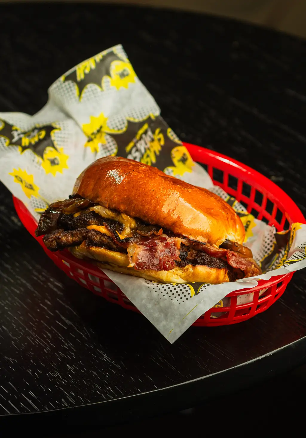 Double beef smash burger with melted vegan cheese, crispy onions, and bacon, held up by gloved hands against a colorful background.
