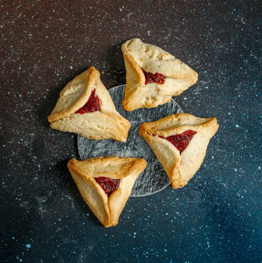 Four triangular hamantaschen cookies filled with red jam, arranged in a circle on a silver plate over a starry dark background.