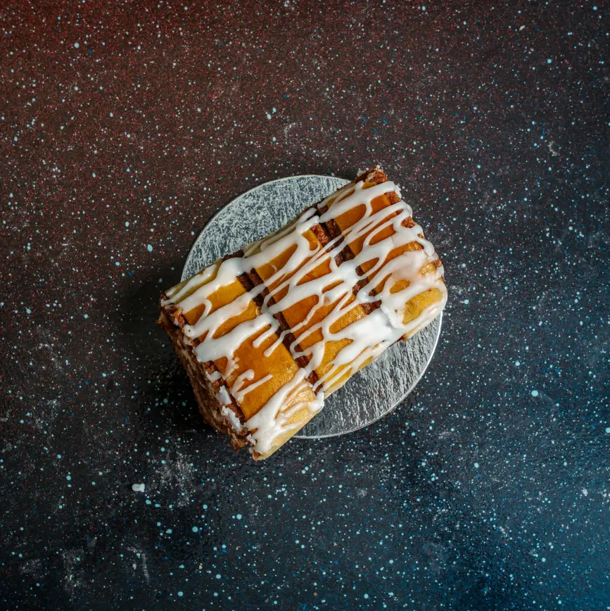 Iced cinnamon babka loaf drizzled with white icing, served on a round silver base against a dark speckled background