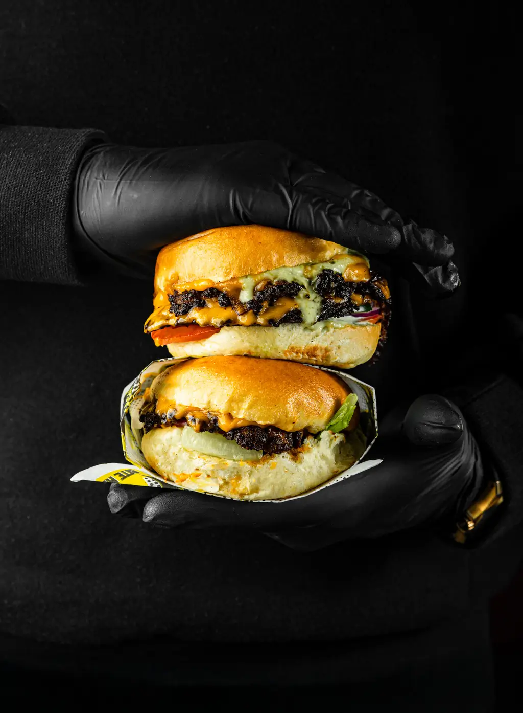 Two stacked smash burgers held in gloved hands, each layered with vegan cheese, grilled onions, lettuce, tomato, and toasted buns.