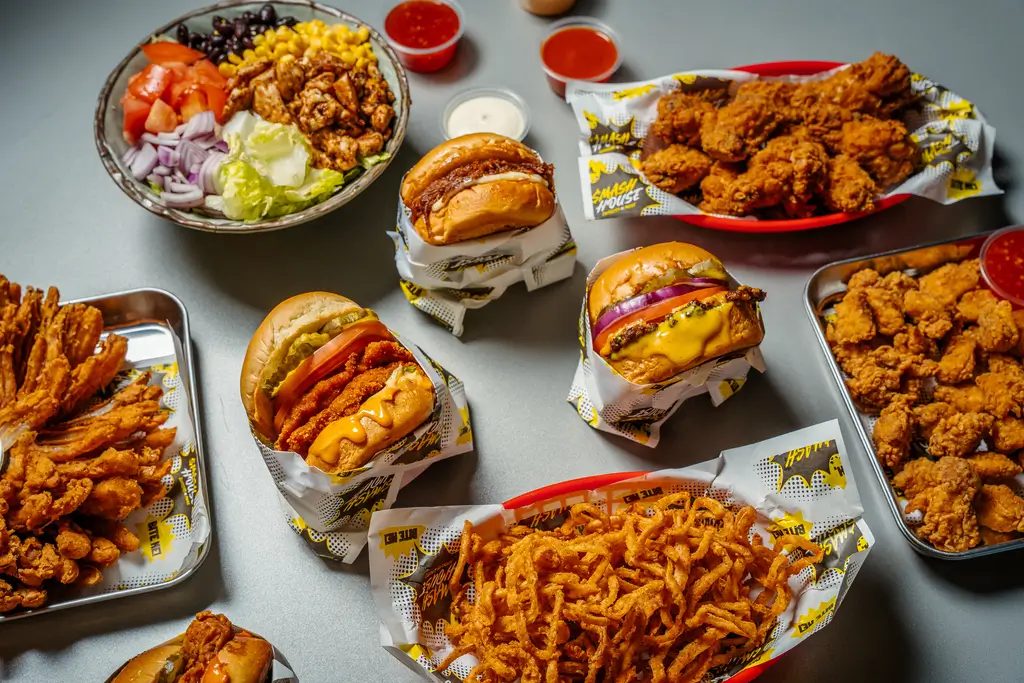 Overhead spread of Smash House burgers, fries, crispy chicken, crispy onions, and a colorful salad bowl, all served with dipping sauces.