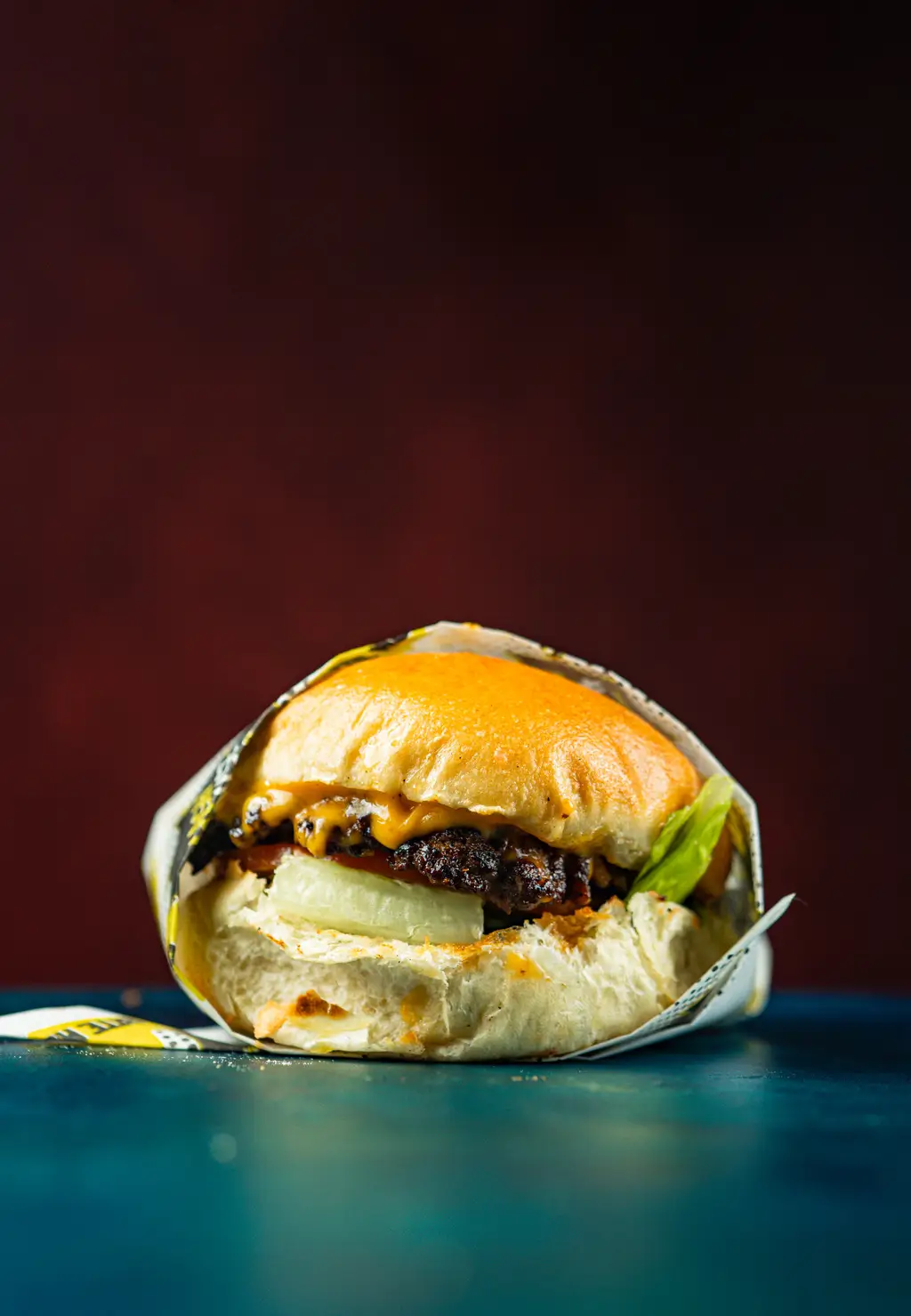 Close-up of a beef smash burger loaded with vegan cheese, grilled onions, green pepper, tomato, and house sauce in a golden toasted bun, wrapped in Smash House paper.