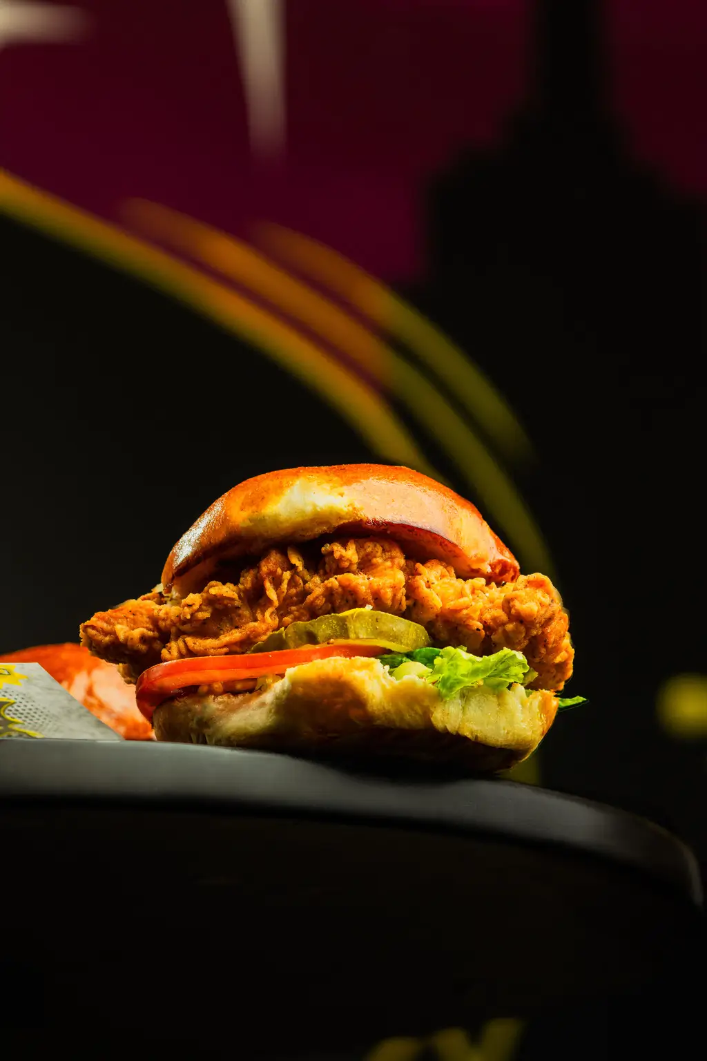 Fried chicken burger stacked with crispy lettuce, tomato, pickles, and a golden fried fillet inside a toasted bun, shot against a dark background with streaks of color.