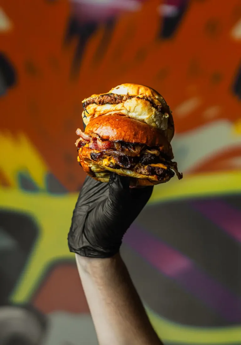 Double stack of smash burgers with vegan cheese and beef bacon held high by a gloved hand, set against a vibrant graffiti-style background
