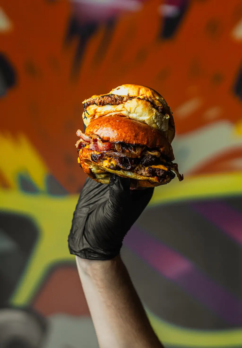 Double stack of smash burgers with vegan cheese and beef bacon held high by a gloved hand, set against a vibrant graffiti-style background