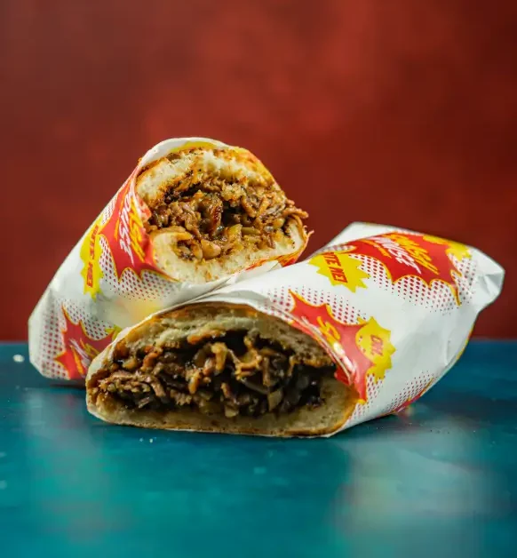 Philly cheesesteak hoagie sliced in half and stacked, filled with thin-cut beef, onions, and sauce, wrapped in branded paper with red comic-style Smash House graphics