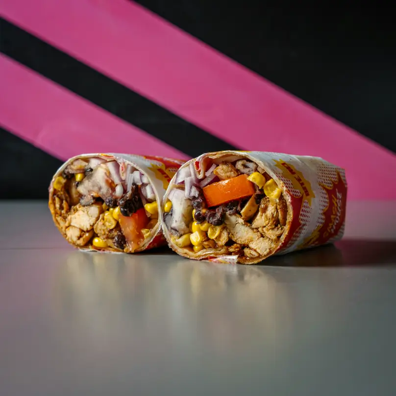 Wrap cut in half, filled with grilled chicken, black beans, corn, tomato, and red onion, wrapped in Smash House packaging and set against a pink-striped wall