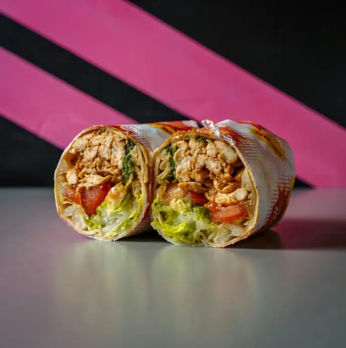 Chicken wrap cut in half and filled with grilled chicken, lettuce, and tomato, wrapped in comic-style Smash House paper against a bold pink and black background