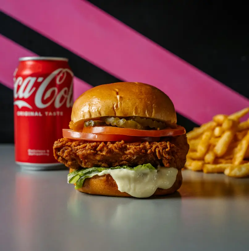 Crispy chicken burger with tomato, lettuce, pickles, and melted vegan cheese on a toasted bun, served with fries and a can of Coca-Cola