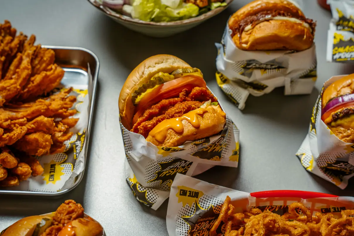 Close-up of a crispy chicken burger layered with pickles, tomato, and melty vegan cheese on a toasted bun, wrapped in Smash House comic-style paper