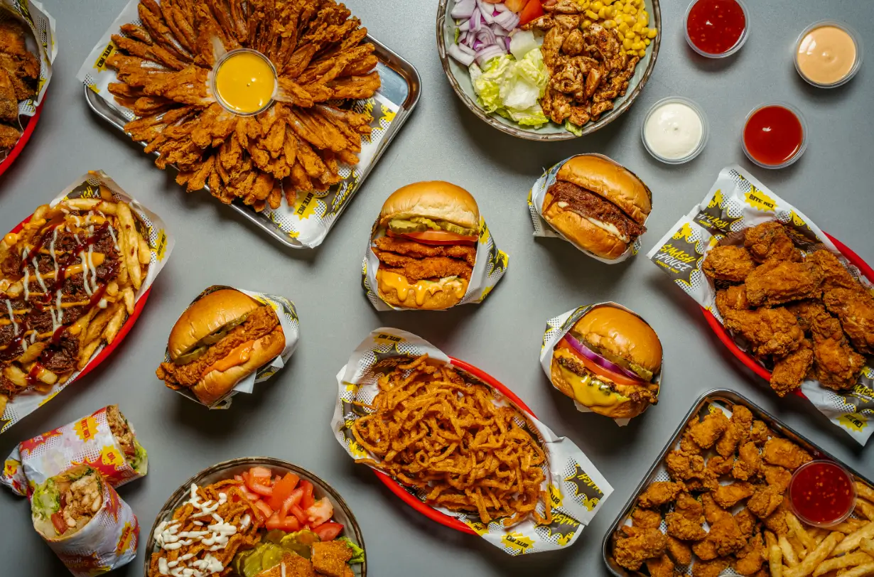 Overhead flat lay of Smash House feast featuring burgers, chicken wings, blooming onion with dip, seasoned fries, chicken wraps, crispy tenders, salads, and dipping sauces