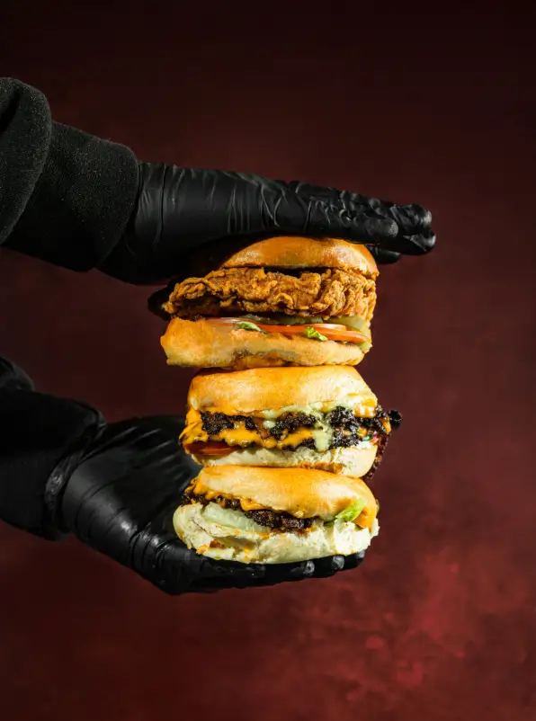 Triple stack of Smash House burgers – one crispy chicken and two beef smash burgers – held in gloved hands against a deep red background