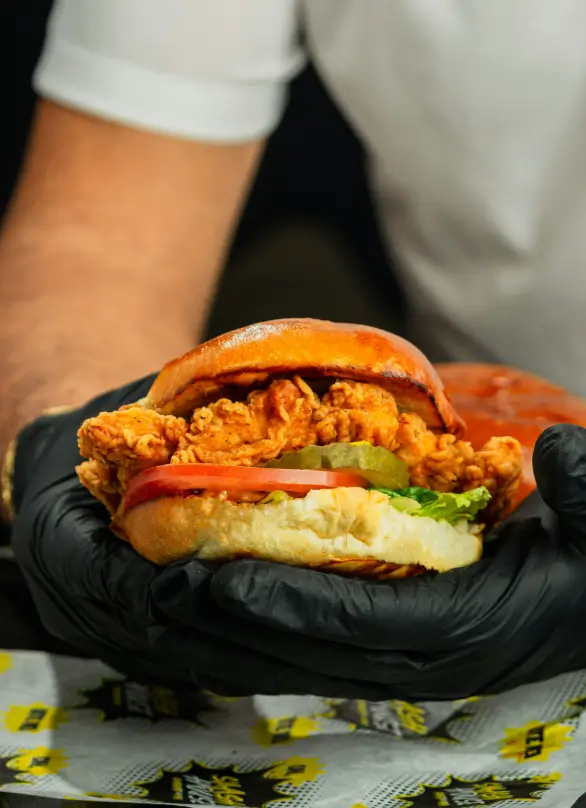Fried chicken burger held in gloved hands, stacked with pickles, lettuce, tomato, and a toasted golden bun, against Smash House branded paper wrap