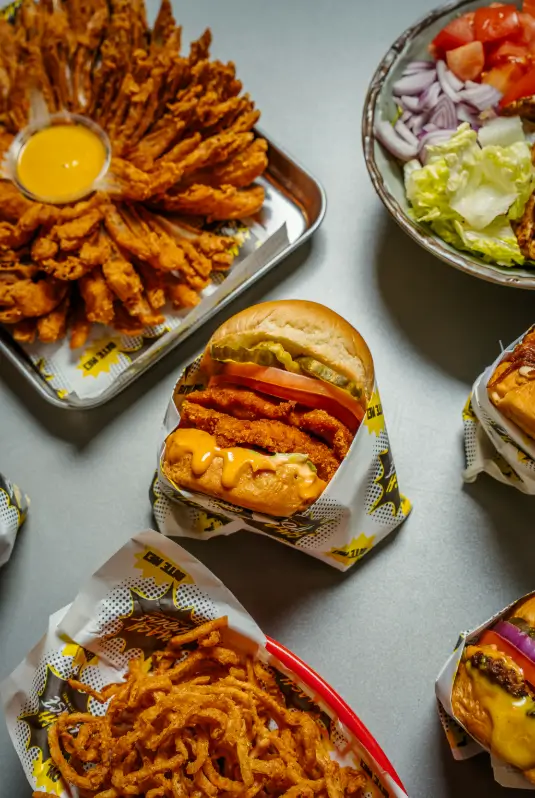 Close-up of a crispy chicken burger layered with pickles, tomato, and melty vegan cheese on a toasted bun, wrapped in Smash House comic-style paper