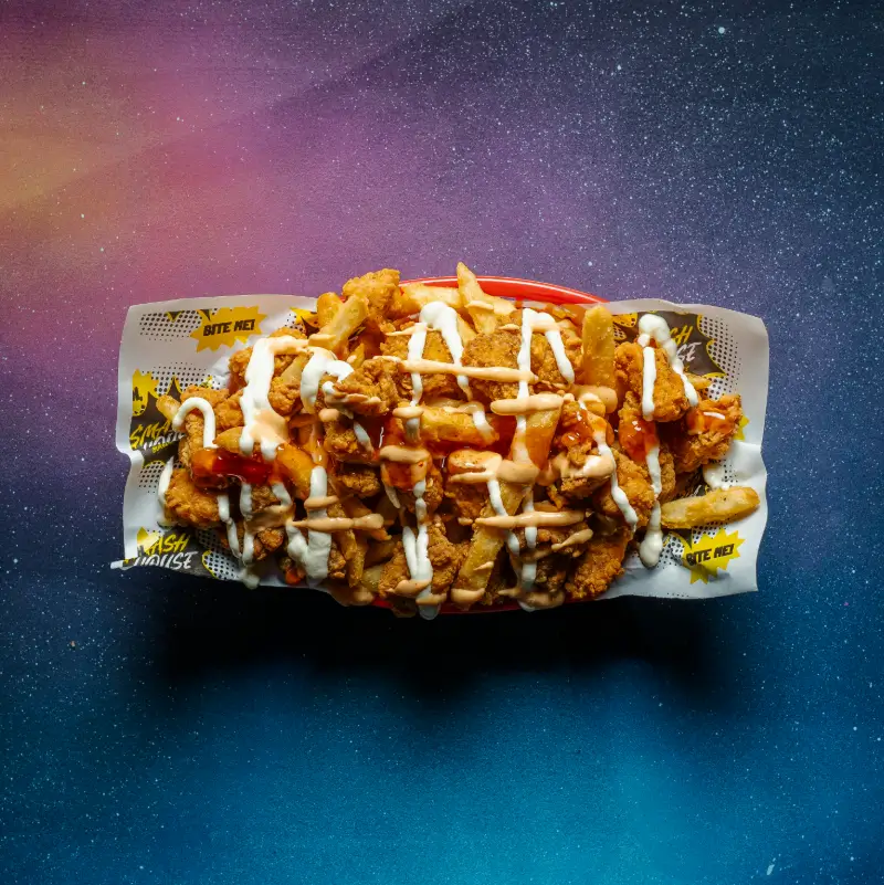 Basket of loaded fries topped with crispy chicken pieces and drizzled with white and orange sauces, served on branded paper reading ‘SMASH HOUSE’ and ‘BITE ME!’ against a vibrant galaxy-themed background