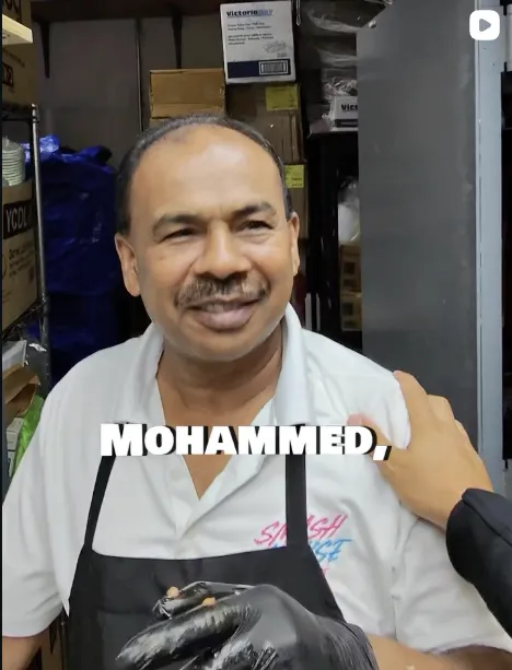 A smiling man wearing a white shirt and black apron with gloves on, standing in a storage area.