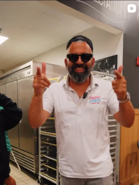 Man with beard and sunglasses giving thumbs up in kitchen setting.