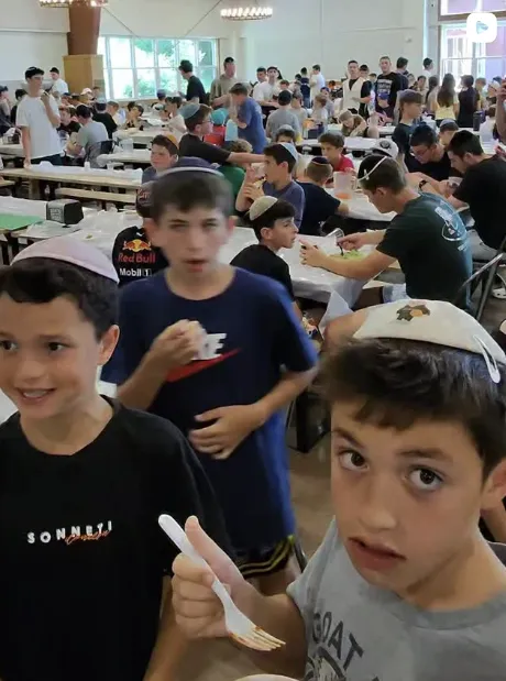 A large group of boys in a dining hall with some wearing kippahs and eating or standing around tables.