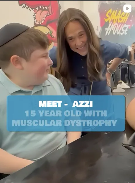 A young boy with muscular dystrophy smiles while sitting next to another person at what appears to be a public event or gathering.